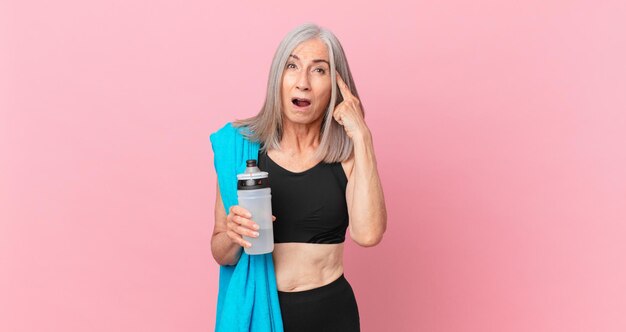 Witte haarvrouw van middelbare leeftijd die verrast kijkt, een nieuwe gedachte, idee of concept realiseert met een handdoek en een waterfles. fitness concept