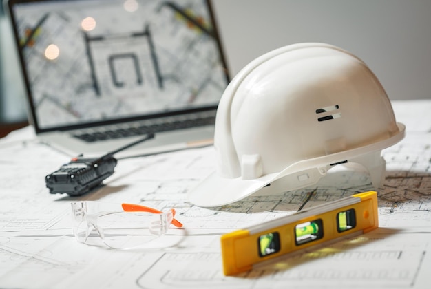 Witte helm, laptop met tekeningen, bril en walkie talkie met en blauwdrukken op een tafel. architectonisch en constructieconcept