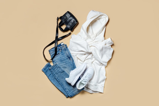 Witte hoody met zak en capuchon met jeans en sneakers op beige achtergrond. Mode sweatshirt, casual jeugdstijl, sport. Stijlvolle herfst- of lentekleding.