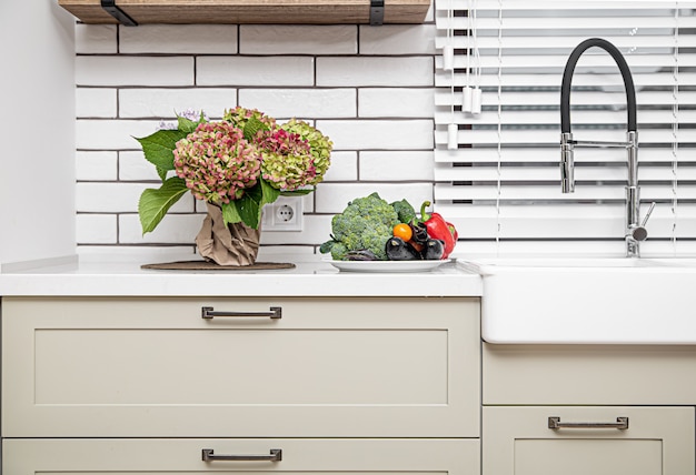 Witte keukenkastjes met metalen handvatten op de deuren bij de wastafel met een bos bloemen en een bordje groenten.