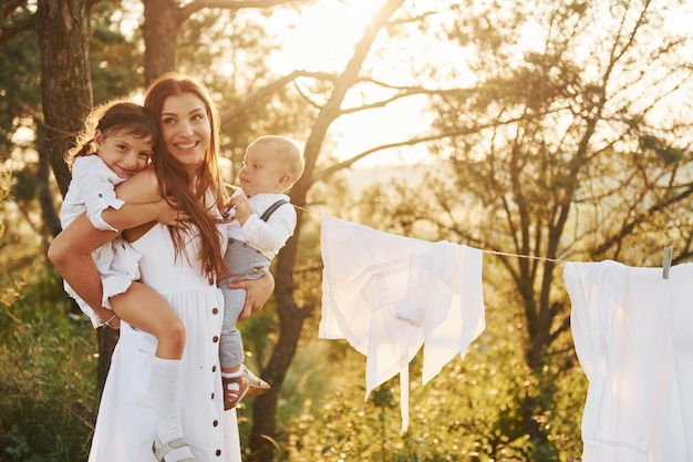 Witte kleren hangen aan het touw om te drogen Jonge moeder met haar dochtertje en zoon is buiten in het bos Mooie zonneschijn