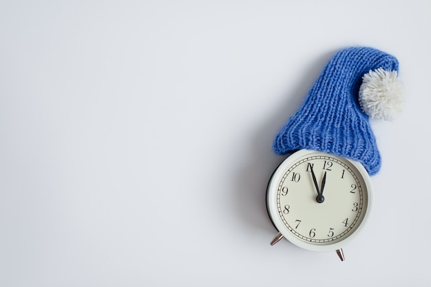 Witte klok in een gebreide blauwe muts Helderwitte achtergrond Vijf minuten voor middernacht Kerstmis