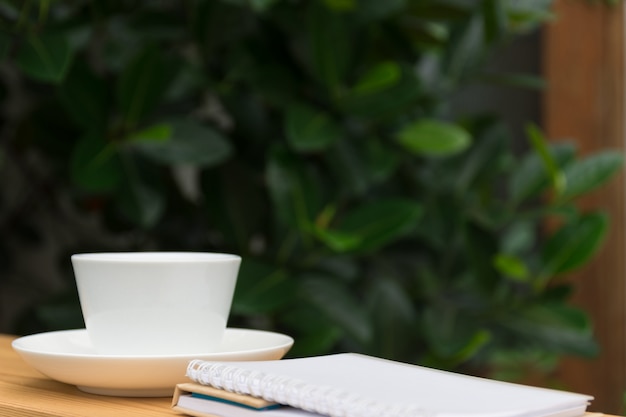 Witte koffiekop en notitieboekje op houten tuin op achtergrond.