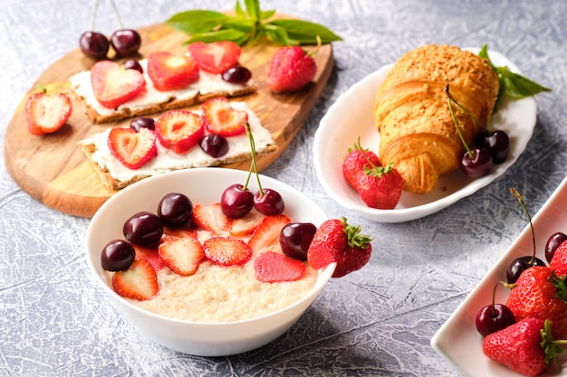 Foto witte kom pap met aardbeien en kersen, croissants en knäckebröd met kaas en aardbeien