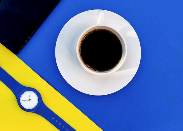 Witte kop koffie op een blauwe achtergrond, blauw handhorloge op gele achtergrond, zwart notitieboekje