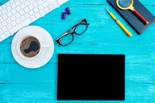 Witte kop koffie, wit toetsenbord, zwarte notebook, zwarte bril en tablet op turquoise houten bureau