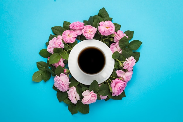 Foto witte kop met zwarte koffie en roze rozen rond rond op een blauwe achtergrond.