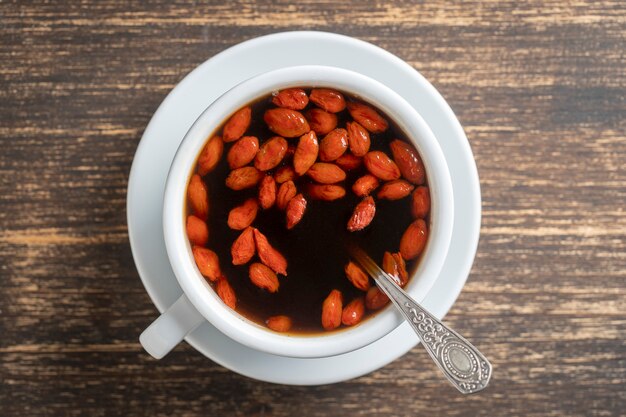 Witte kopje verse ochtend goji-koffie op houten tafel, bovenaanzicht, close-up. Zwarte hete koffie met hele rode gojibessen