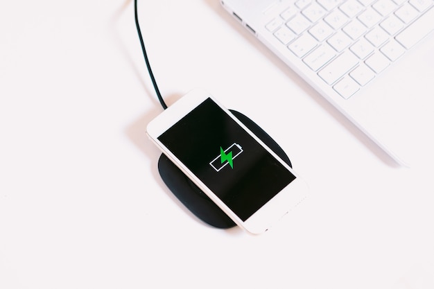 Witte mobiele smartphone met het logo van een batterij met groene bliksem op het scherm, opladen op een oplader zonder kabel naast de laptop op een witte werktafel