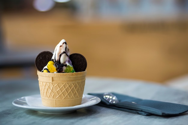 Witte plaat met ijs dessert in wafel kopje met chocolade koekjes en creatieve decoratie topping op wazig kleurrijk interieur.