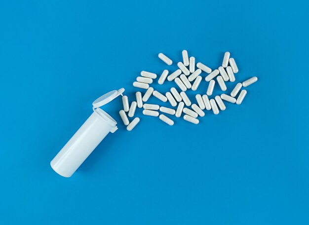 Witte plastik medische fles en verspreide pillencapsules op een blauwe achtergrond met exemplaarruimte.