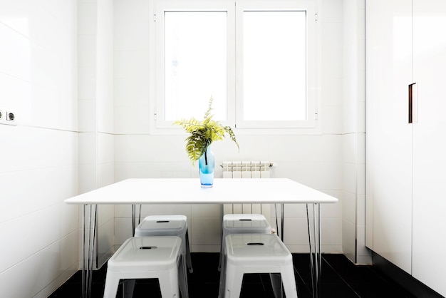 Witte rechthoekige houten eettafel met blauw glazen vaas met bloemen en wit metalen krukken onder een aluminium raam