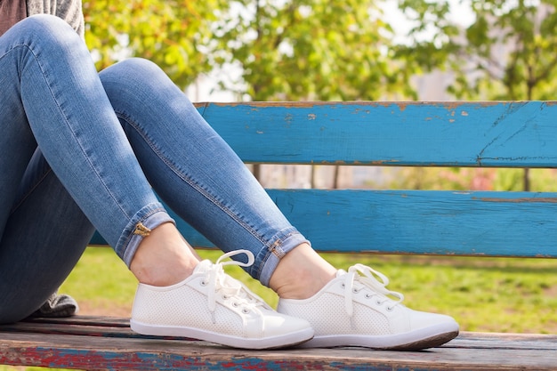 Witte sneakers op vrouwelijke benen in spijkerbroek op een blauwe bank en een groene plant achtergrond.