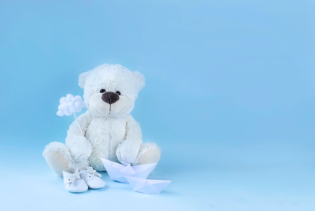 Witte teddybeer met een kleine wolk, babyslofjes en twee papieren bootjes op een lichtblauwe ondergrond