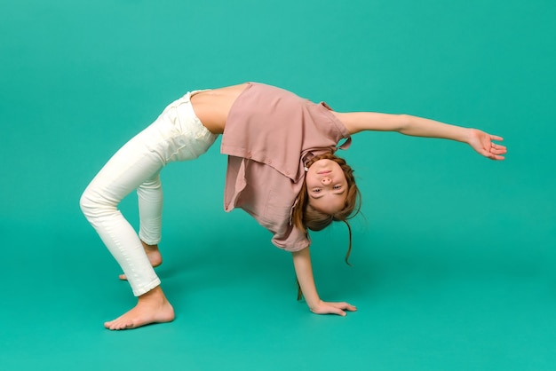 Witte tienermeisje in witte broek en een roze shirt staat in de pose van een brug op een groen oppervlak, acrobaat