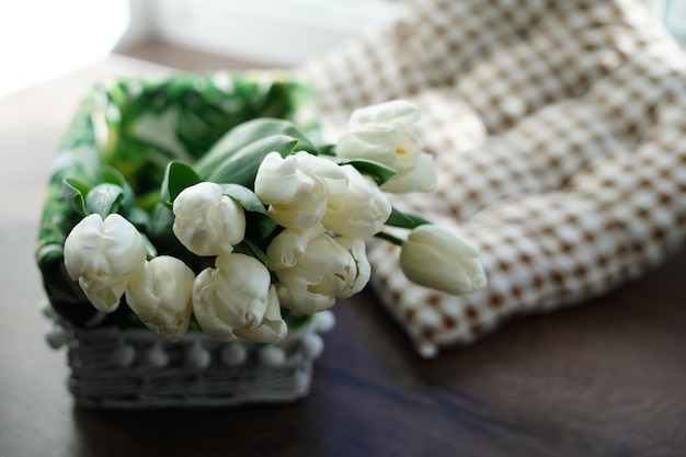 Witte tulpen liggen in een decoratieve witte rieten mand op een houten tafel bij het raam