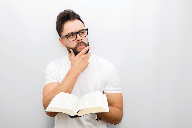 Wize and thoughtful young man in glasses stand and hold opened book in hands. He looks to side. Guy holds hand on chin.