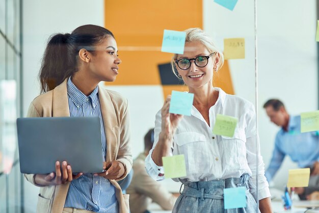 Wo happy female coworkers putting colorful sticky notes on a glass wall and smiling while standing