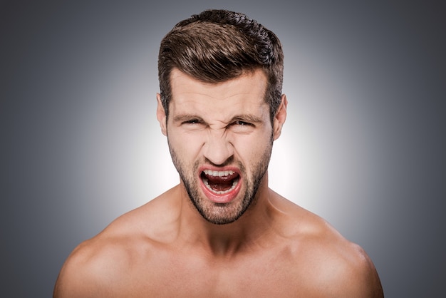 Woedend mens. Portret van een woedende jonge shirtloze man die naar de camera kijkt en zijn mond openhoudt terwijl hij tegen een grijze achtergrond staat