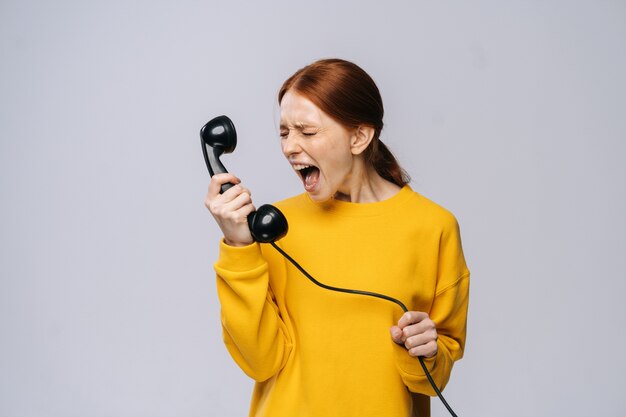 Woedende boze jonge vrouw in stijlvolle gele trui praten over retro telefoon en schreeuwen in handset