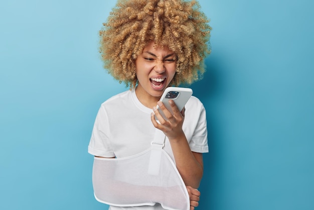 Woedende vrouw schreeuwt in de telefoon drukt woede uit schreeuwt luid heeft gebroken arm in draagdoek draagt casual wit t-shirt geïsoleerd op blauwe achtergrond boos om spam-oproep moe van vervelende robocalls