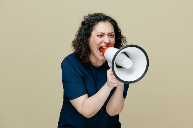 Woedende vrouw van middelbare leeftijd draagt t-shirt met luidspreker met beide handen en kijkt ernaar en schreeuwt hardop in luidspreker geïsoleerd op olijfgroene achtergrond