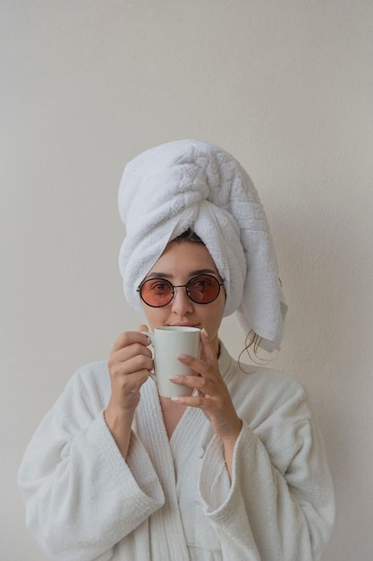 Una donna con gli occhiali in accappatoio e asciugamano in hotel beve caffè da una tazza bianca. fotografia gioiosa di stile di vita