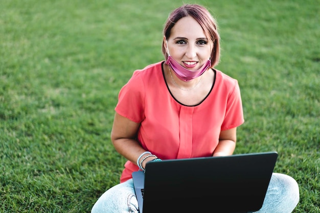 Donna di 40 anni che lavora all'aperto con il laptop durante l'epidemia di coronavirus imprenditrice sorridente seduta sul prato con un personal computer mentre guarda la fotocamera concetto di lavoro a distanza nella natura