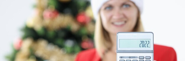 Woman accountant in santa claus hat holds calculator on background of christmas tree summing up