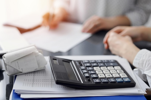 Woman accountant using a pen and laptop computer while counting and discussing taxes with a client, focus on the calculator. Business audit and finance concepts.