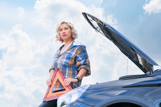 A woman against the background of a broken car and a cloudy sky holds a triangular emergency stop sign in her hand while standing at the open hood