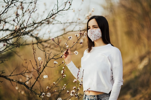 春の開花の木を背景にした女性は、彼女の顔に医療用マスクを示しています。