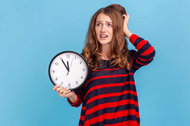 Woman anxiously looking aside holding clock in hand frustrated with deadline being late