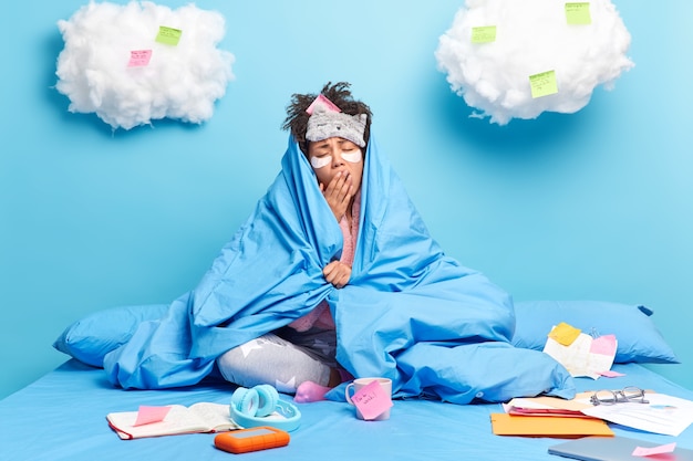 woman applies beauty patches yawns as works late hours wrapped in blanket prepares for final exams