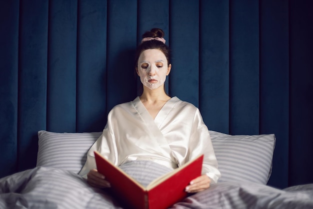 Foto la donna applica una maschera di pulizia sul viso effetto ringiovanimento si siede sul letto e legge il libro rosso
