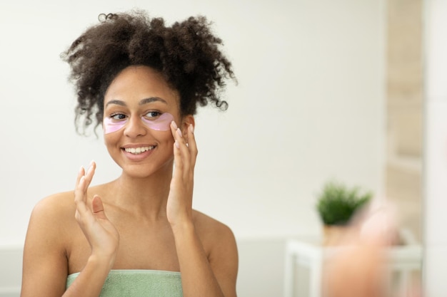Foto una donna si applica i cerotti sotto gli occhi, prendendosi cura della pelle del viso, in piedi vicino allo specchio