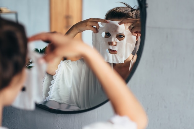 여자는 거울을보고 그녀의 얼굴에 시트 마스크를 적용