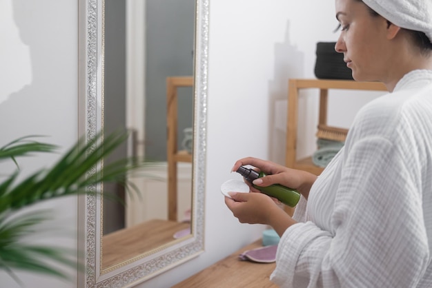 Woman applying cleaning spray on cotton disk near mirror in bathroom lady doing skincare routine