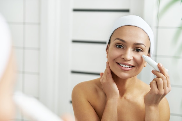 The woman applying cream under the eyes while looking at herself through the mirror