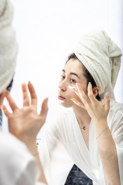 Woman applying cream on her face in front of the bathroom mirror