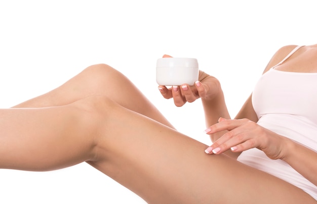 Woman applying cream onto her legs on white wall. Skin care and health concept.