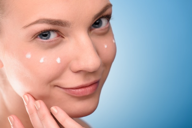 Woman applying lifting cream on face over blue background.