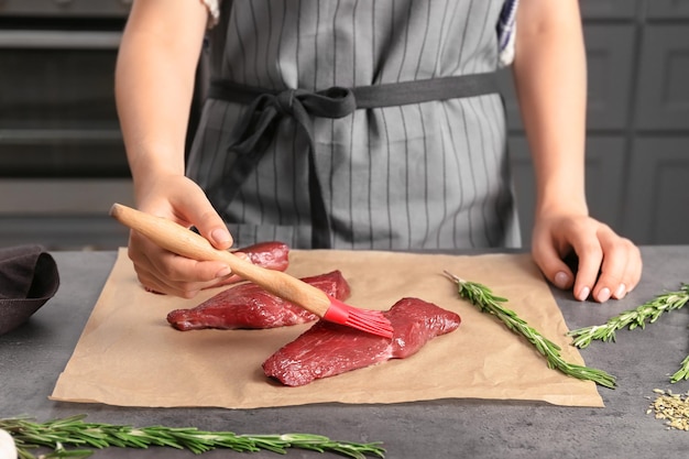 Woman applying oil onto raw steak with silicone brush in kitchen