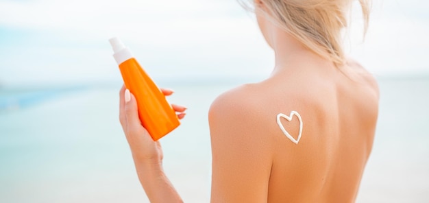 Woman applying sun cream on tanned body In form heart love to protect her skin Girl using sunscreen Female holding suntan lotion moisturizing sunblock on sunny day with blurry sea in the background