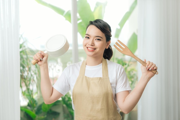 Woman in apron in hand frying pan and kitchen spatula near big window