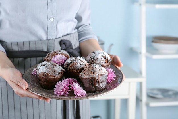 Woman in apron holding plate with chocolate muffins on kitchen closeup