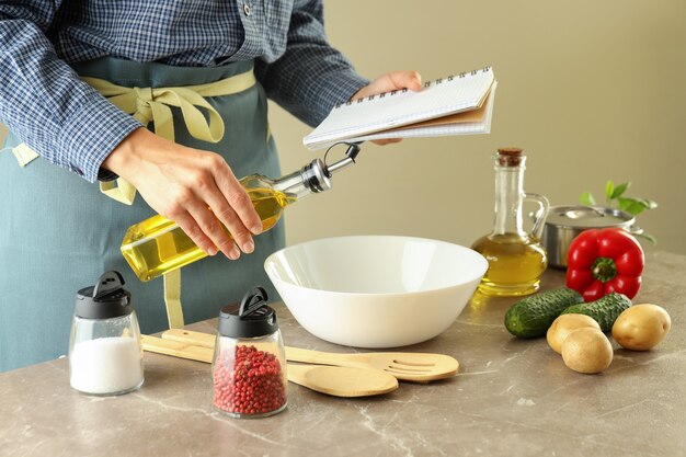 A woman in an apron holds a recipe book and a bottle of oil and cooks