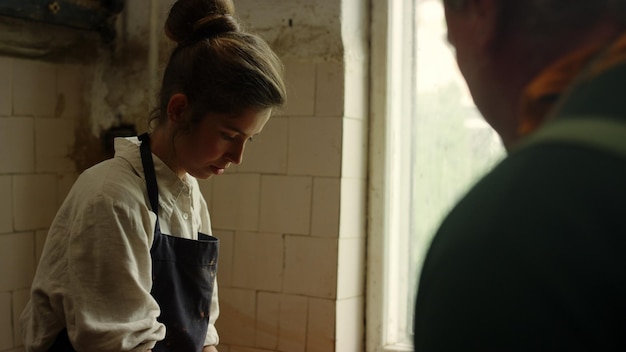 A woman in an apron is standing in front of a window with a man in the background.