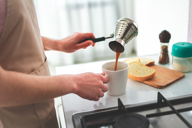 自宅のキッチンで朝食にcezveから熱いおいしいトルココーヒーを注ぐエプロンの女性。