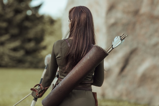 Woman archer with arrows on her back, stands with her back to the viewer on a blurred background, close-up